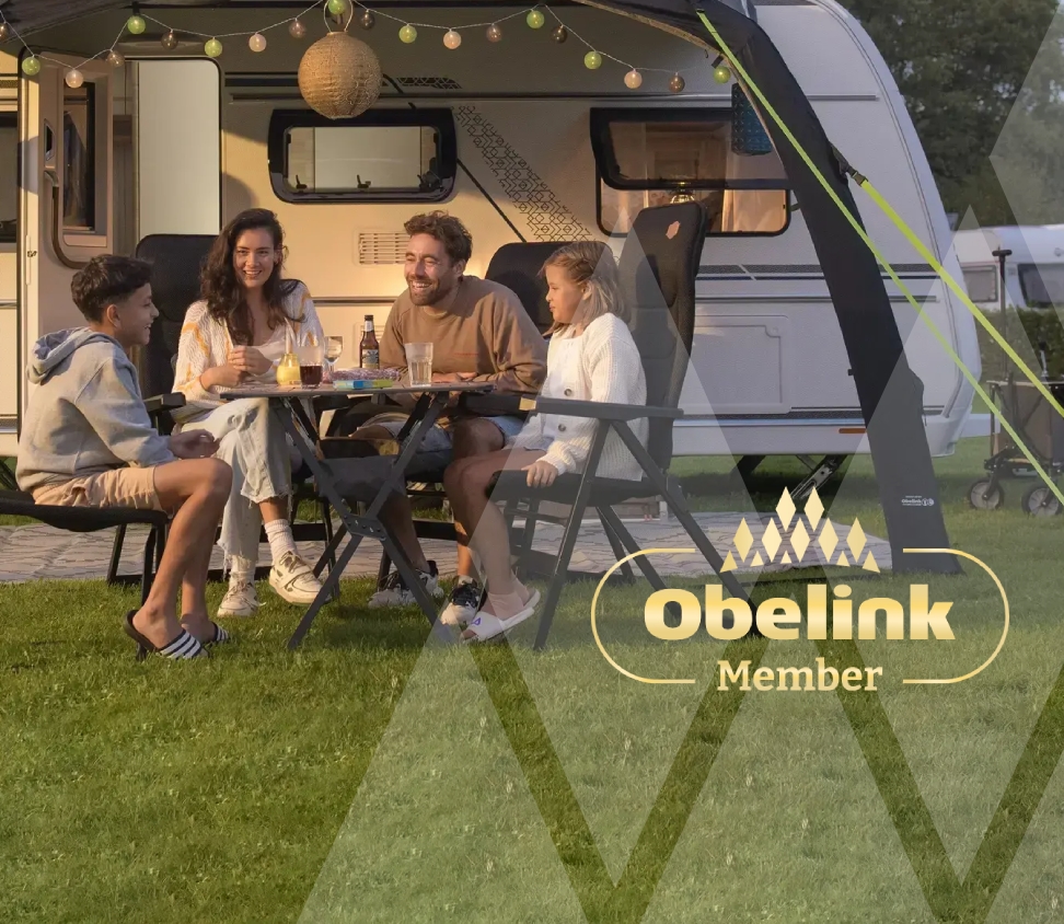 Caravan met luifel versierd met sfeerverlichting, mensen genieten samen aan een kampeertafel in de avondschemering op een grasveld
