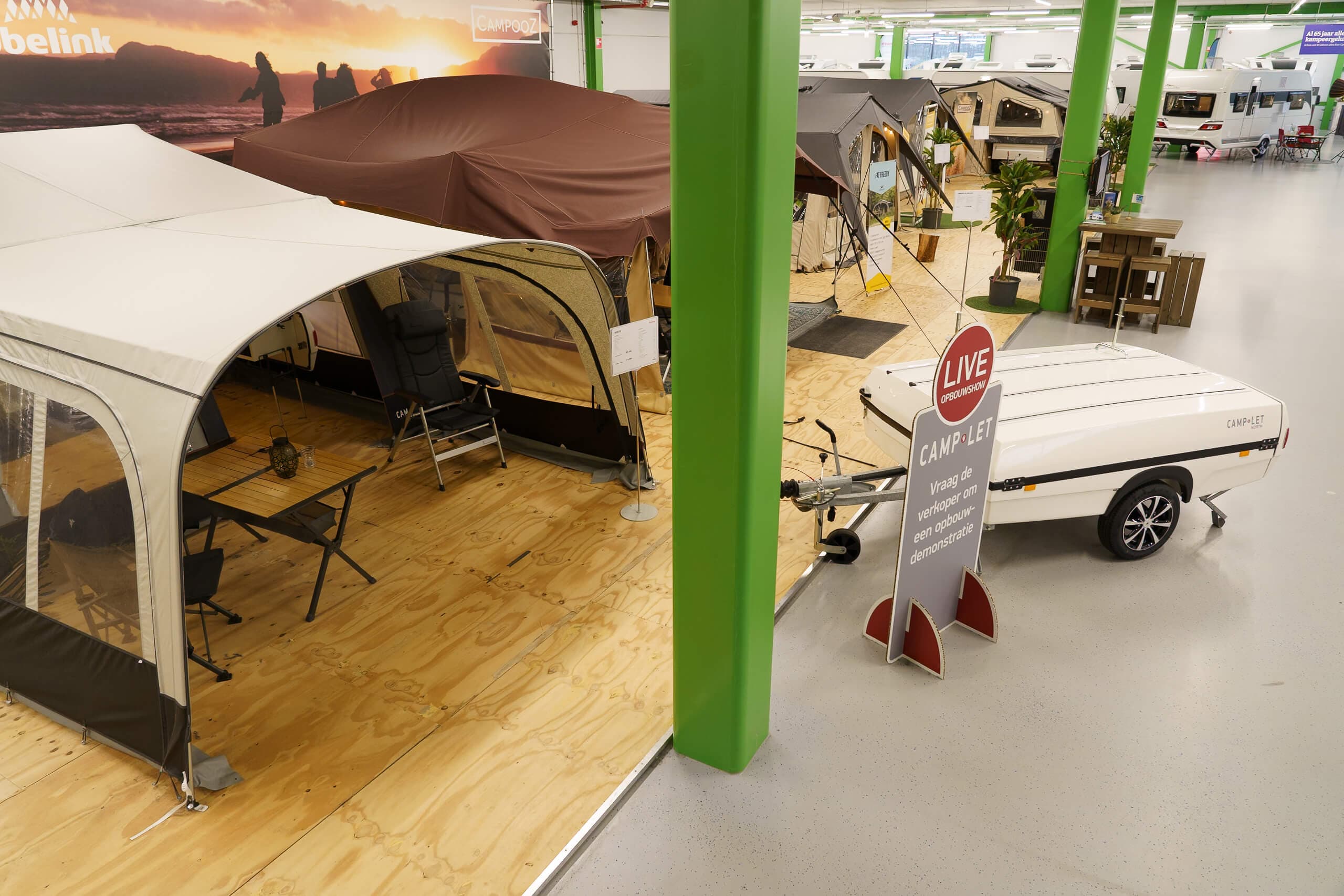 Showroom met verschillende vouwwagens, tenten en caravans op houten vloer met Camp-let aanhangwagen en 'LIVE OPBOUWSHOW' bord op de voorgrond