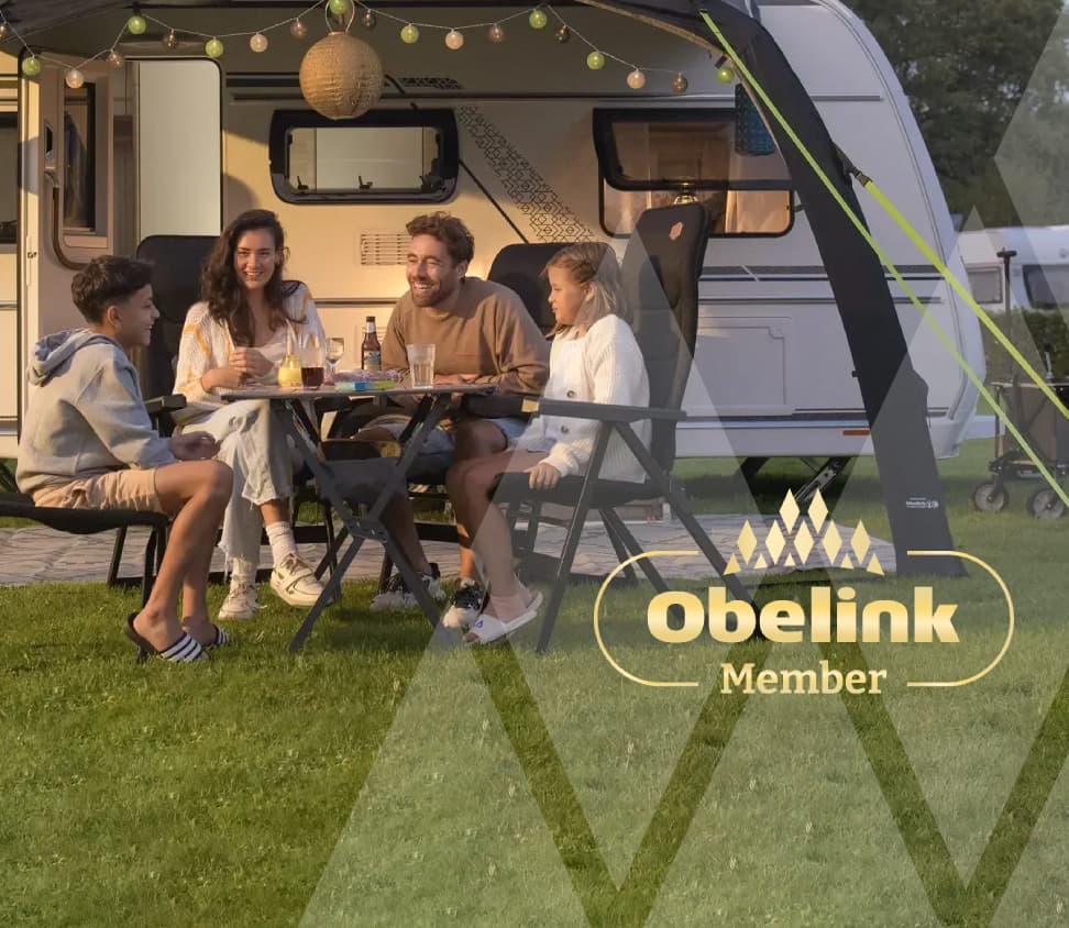 Sparen voor korting Caravan met luifel versierd met sfeerverlichting, mensen genieten samen aan een kampeertafel in de avondschemering op een grasveld