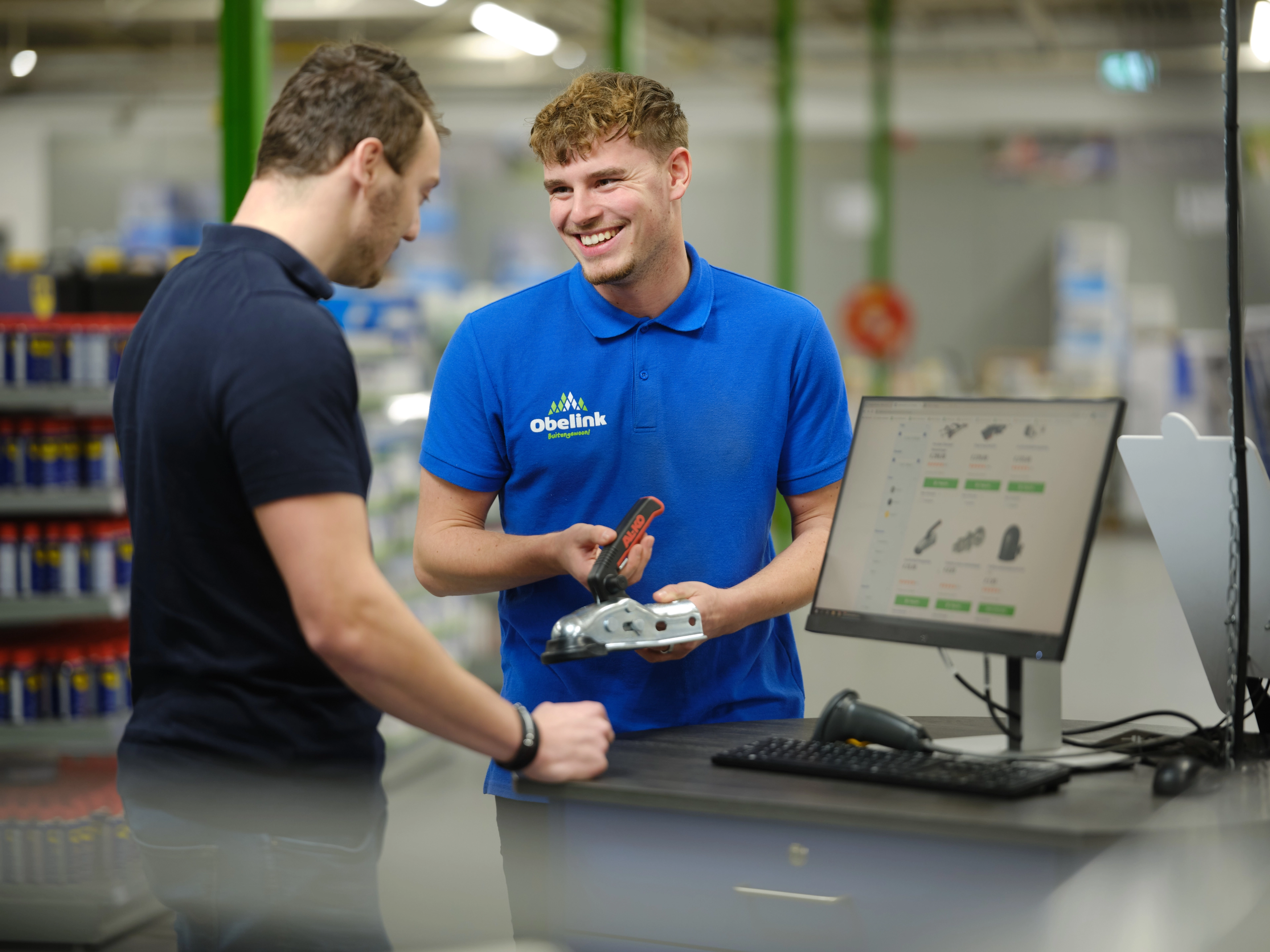 Kom je er niet helemaal uit? Medewerker in blauw Obelink poloshirt toont een metalen bevestigingsonderdeel aan klant bij verkoopbalie met computerscherm waarop producten zichtbaar zijn.