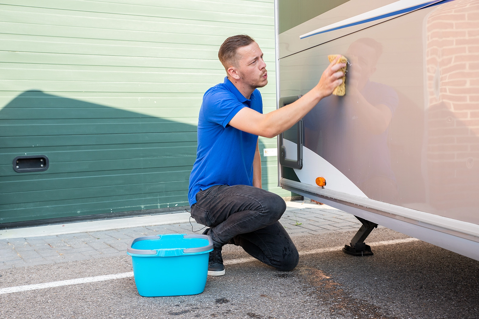 caravan schoonmaken met spons Man in blauw t-shirt reinigt zijkant van caravan met spons, blauwe emmer water ernaast op oprit