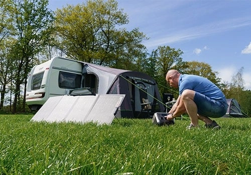 man zonnepanelen aanzetten op de camping Man plaatst zonnepanelen voor een voortent bij een caravan op een grasveld omringd door bomen onder een blauwe hemel