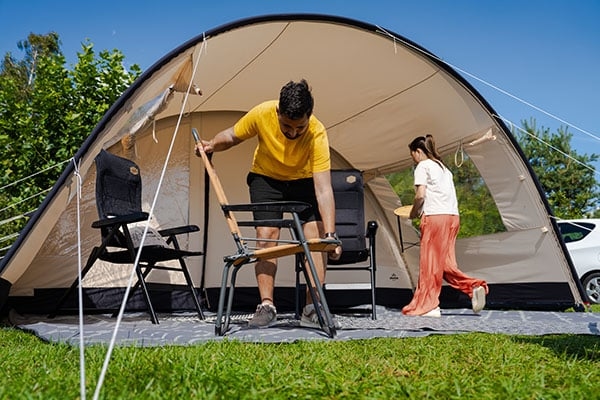 Man vouwt stoel uit voor tent Ruime koepeltent met voortent op grasveld, twee personen zetten kampeerstoelen en tafel klaar onder zonnig blauwe hemel