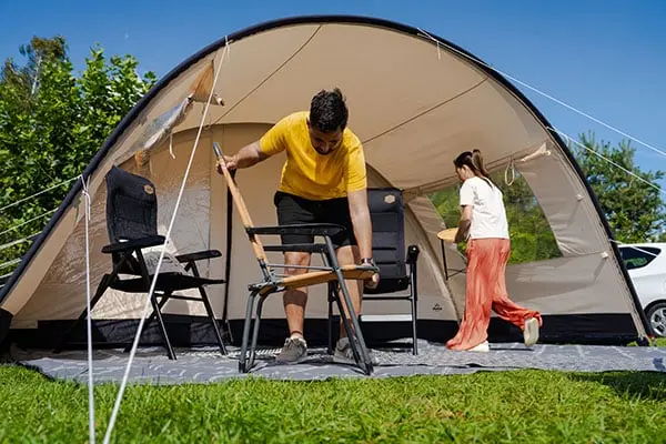 Man vouwt stoel uit voor tent Ruime koepeltent met voortent op grasveld, twee personen zetten kampeerstoelen en tafel klaar onder zonnig blauwe hemel
