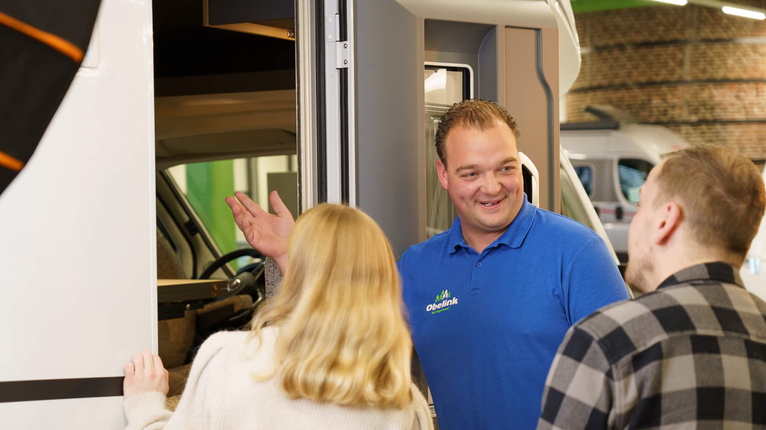 verkoper bij caravan met 2 klanten Medewerker in blauw Obelink poloshirt toont een camper aan klanten in de showroom